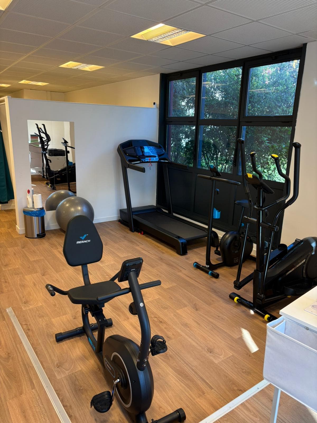 Salle de rééducation lumineuse avec tapis de course, vélo et vélo elliptique devant de grandes fenêtres